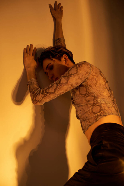 Een jonge homo in stijlvolle kleding leunt tegen een muur met uitgestrekte armen, belichaamt kracht en trots. - Foto, afbeelding