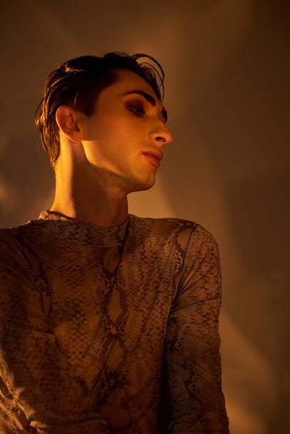 Ein junger queerer Mann im Schlangenhaut-Hemd steht selbstbewusst in einem schwach beleuchteten Raum.. - Foto, Bild