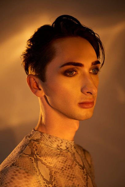 Ένας νεαρός queer άντρας σε ένα φόρεμα δέρμα φιδιού στέκεται με χάρη μπροστά από ένα φωτεινό φως, αποπνέοντας υπερηφάνεια και στυλ. - Φωτογραφία, εικόνα