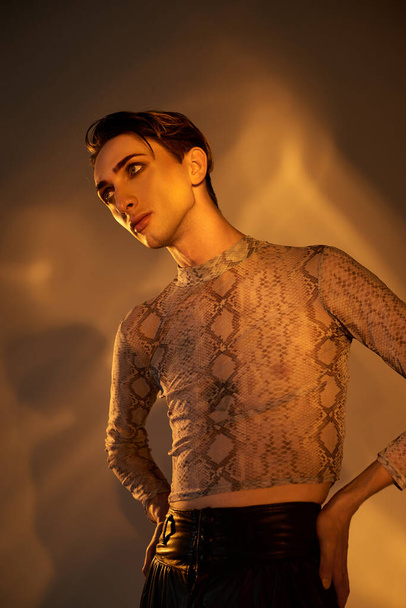 Ένας νεαρός queer άνδρας με αυτοπεποίθηση θέτει μπροστά από ένα φωτεινό φως σε κομψό τοπ και δερμάτινο παντελόνι. - Φωτογραφία, εικόνα