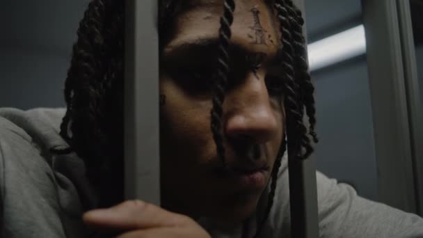 Yüzünde dövmeler olan depresif Afro-Amerikan genç suçlu hapishane hücresinde duruyor, metal parmaklıkları var. Genç mahkûm, hapiste işlediği suçtan hapis cezasını çekiyor. Islahevi. Kapat.. - Video, Çekim