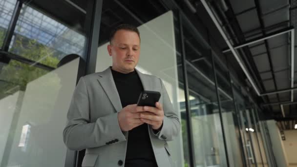 With a smartphone in hand, an executive stands absorbed in the digital world, framed by the geometric elegance of a modern office atrium. - Footage, Video