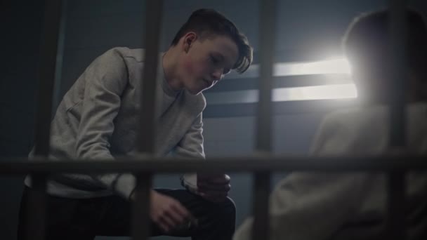 Caucasian teenage prisoner plays cards in prison cell with other inmates. Young criminals serve imprisonment terms in jail. Juvenile detention center or correctional facility. View through metal bars. - Footage, Video