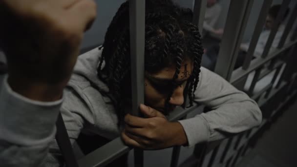 Upset African American teenage prisoner stands in prison cell in jail, holds metal bars. Young inmates play cards on bed in the background. Youth detention center or correctional facility. High angle. - Footage, Video