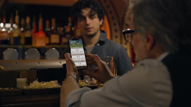 Mature sports fan bets on football match online using mobile phone, talks with bartender and friend. Man checks game score and bookmaker ratings in app sitting at bar counter in pub. Gambling concept. - Footage, Video