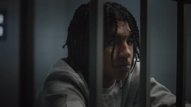 Dühös afro-amerikai tinédzser fogoly arctetoválással a börtön cellájában áll és a kamerába néz. Fiatal bűnözőt börtönbüntetésre ítéltek. Fiatalkorúak börtönében. Portré. - Felvétel, videó