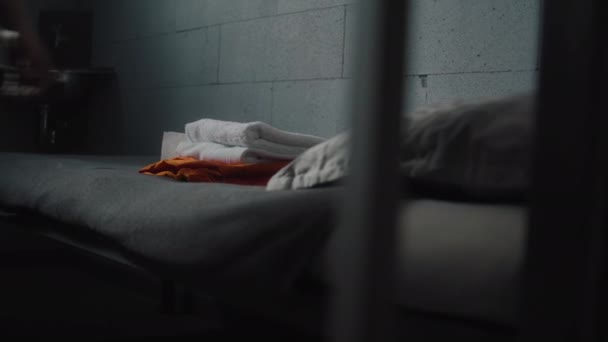 Prisoner puts orange uniform and bathroom accessories on the bed in prison cell. Criminal, inmate serves imprisonment term in jail. Detention center or correctional facility. View through metal bars. - Footage, Video