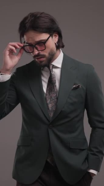 κάθετο βίντεο με ελκυστικό νεαρό άνδρα σε κομψό κοστούμι περπάτημα με το χέρι στην τσέπη, ρυθμίζοντας γυαλιά ηλίου και κοστούμι μπροστά από το γκρι φόντο - Πλάνα, βίντεο