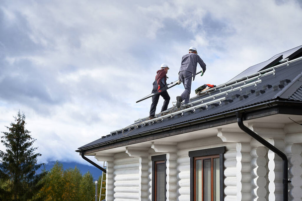 Electriciens installant un système de panneaux solaires sur le toit de la maison. Hommes travailleurs dans les casques transportant module solaire photovoltaïque à l'extérieur. Concept d'énergies alternatives et renouvelables. - Photo, image