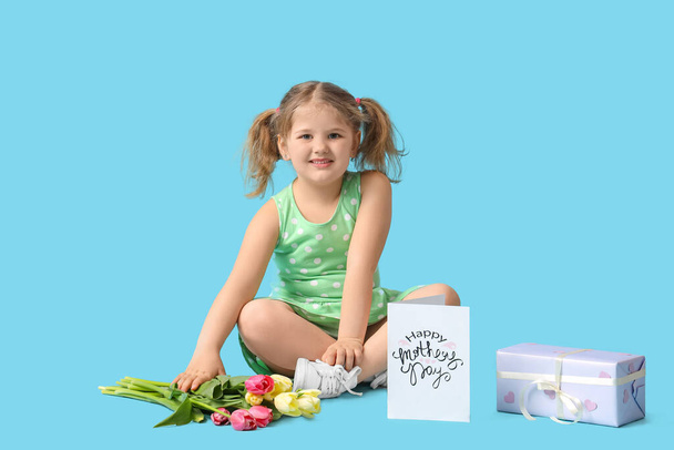 Little girl sitting with bouquet of tulips , gift box and greeting card on blue background. Mothers Day concept - Photo, Image