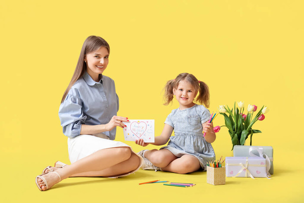 Mom with her little daughter sitting on floor with bouquet of tulips and greeting card on yellow background. Mothers Day concept - Photo, Image