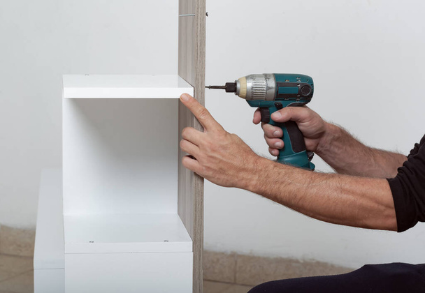 Un homme assemblant une étagère blanche. Une perceuse électrique est utilisée pour fixer une planche de bois à l'étagère. Les mains de mans sont visibles, indiquant le processus d'assemblage. L'étagère est contre un mur blanc. - Photo, image
