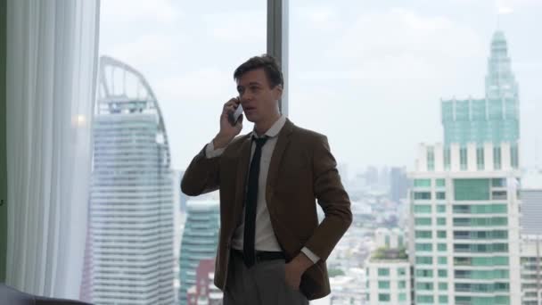 Uomo d'affari in piedi in ufficio ornato a parlare al telefono con il college su sfondo finestra skyline paesaggio urbano. Determinazione e ambizione guidano la carriera aziendale verso un futuro luminoso - Filmati, video