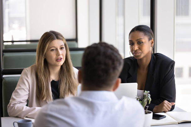 Multiethnic female HR team talking to male job candidate at interview meeting in coworking space. Caucasian and African businesswomen speaking to male colleague at workplace table - Photo, image