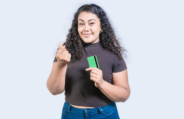 Привлекательная девушка, держащая кредитную карту, зарабатывает деньги жестом с изолированными пальцами, глядя в камеру - Фото, изображение