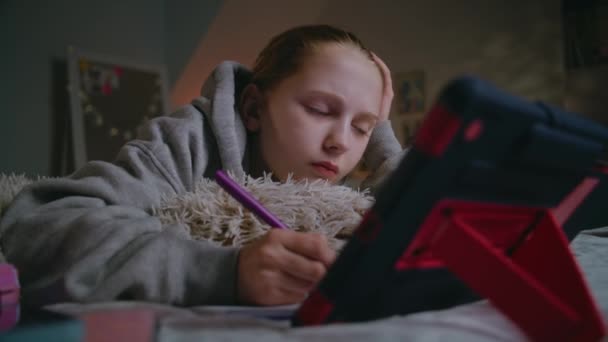 Jong blank meisje ligt thuis op bed, schrijft in notitieboekje en doet online schoollessen met behulp van digitale tablet. Tienermeisje brengt overdag tijd door en studeert in gezellige slaapkamer. Levensstijl concept. - Video