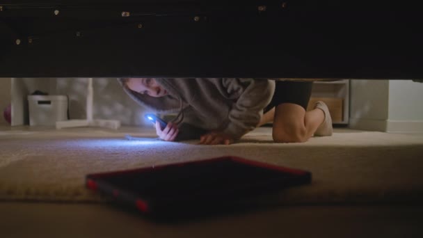 Jong meisje komt naar de grote licht slaapkamer met zaklamp op haar mobiele telefoon. Blanke tiener kruipt onder bed, vindt digitale tablet computer en gebruikt het. Schieten onder het bed. Levensstijl concept. - Video