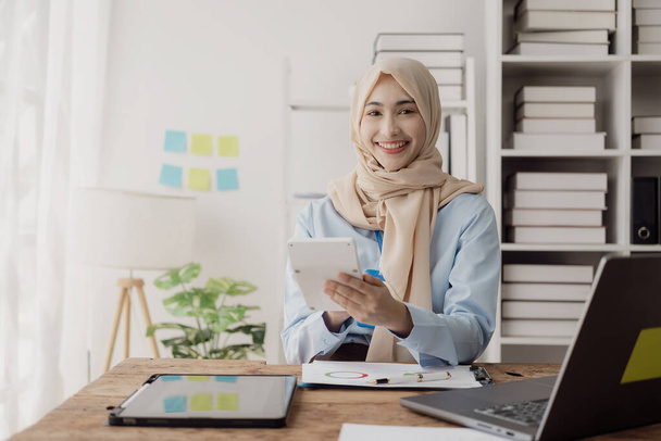 Asyalı Müslüman iş kadını hesap makinesi kullanır ve masadaki dizüstü bilgisayarlı ve mali belgeli, vergi, muhasebe, finansal hesaplı notlar yazar. - Fotoğraf, Görsel