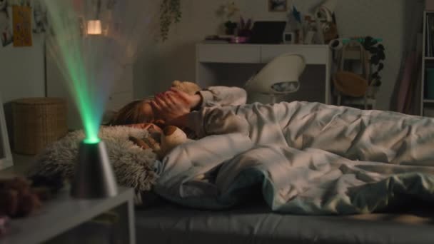 Young girl lies on bed under blanket with plush toys and rests in her room in the evening. Schoolgirl wakes up, looks around and uses phone late night. Comfortable home with cozy and stylish interior. - Footage, Video