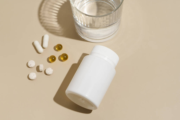 Fehér mockup üres üveg és kapszula tabletták bézs izolált háttér pohár vízzel. Egészségügyi ellátás, betegségkezelés, vitaminok szépségápolási koncepcióhoz. - Fotó, kép