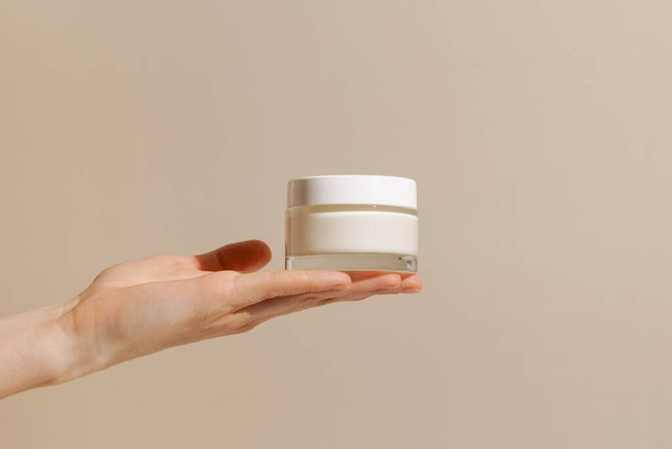 Female hand holding white glass mockup jar of face cream on beige isolated background. Concept of beauty products, skin care, nutrition and hydration - Photo, Image