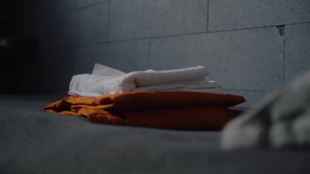 Lähikuva vangin asettamalla oranssi vankilan univormu ja kylpyhuoneen tarvikkeet sängylle. Syyllinen tai vanki istuu tuomiota rikoksesta vankilassa. Säilöönottokeskus tai vankila. Oikeusjärjestelmä. - Materiaali, video