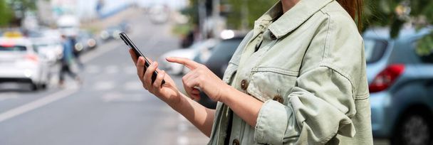 Nainen kaupunkilainen seisoo tien vieressä kaupungissa ja käyttää älypuhelintaan. Autonvuokraus, taksi, julkinen liikenne. - Valokuva, kuva