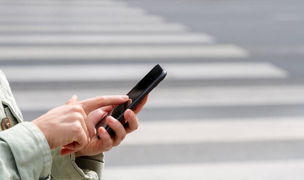 Un smartphone dans les mains d'une femme devant un passage piétonnier dans la ville.  - Photo, image