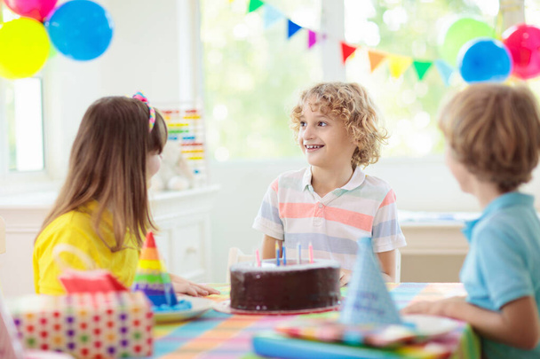 Kids birthday party. Child blowing candles on cake and opening presents. Pastel rainbow theme celebration. Family celebrating at home. Boy opening gifts, eating cakes. Sweets for children. - Photo, Image