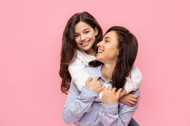 Frau Mutter hat Spaß mit ihrer niedlichen Tochter, Mädchen umarmt Mutter von hinten, posiert zusammen auf pastellrosa Wand Hintergrundstudio - Foto, Bild