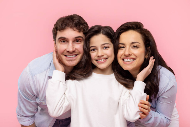 Retrato de familia joven de tres, padre, madre e hija bonita sonriendo a la cámara, niña tocando las mejillas de sus padres, fondo rosa - Foto, imagen
