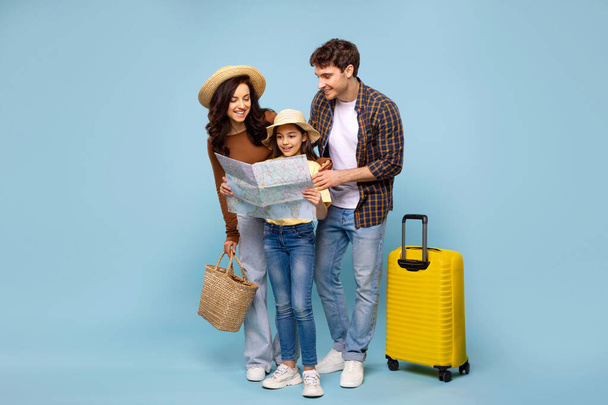 Happy family with daughter looking at map, planning travel, standing with suitcase on blue studio background, ready for summer vacation - Photo, Image