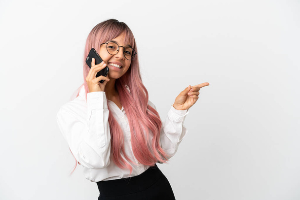 Молодий бізнес змішаної раси жінка з рожевим волоссям тримає мобільний ізольований на рожевому фоні вказуючи пальцем на бік - Фото, зображення