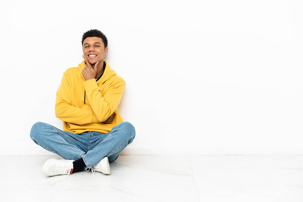 Jeune homme afro-américain assis sur le sol isolé sur fond blanc souriant - Photo, image