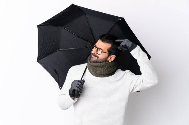 Caucásico hombre guapo con barba sosteniendo un paraguas sobre la pared blanca aislada que tiene dudas mientras se rasca la cabeza - Foto, imagen