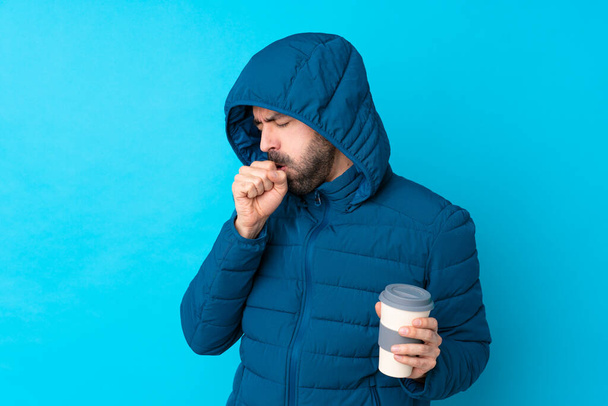 Mężczyzna noszący zimową kurtkę i trzymający kawę na wynos na odizolowanym niebieskim tle cierpi z powodu kaszlu i złego samopoczucia. - Zdjęcie, obraz