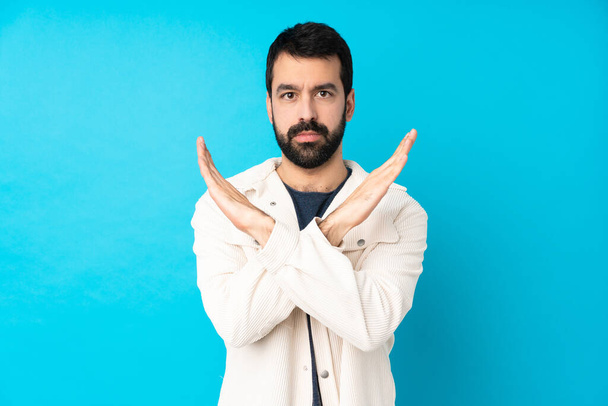Young handsome man with white corduroy jacket over isolated blue background making NO gesture - Photo, Image
