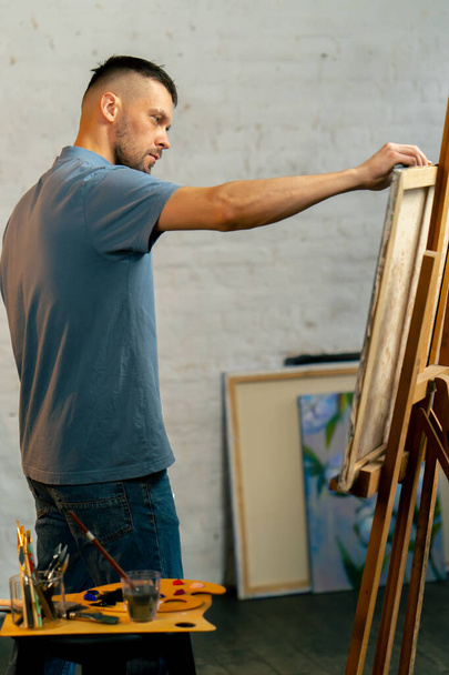 σε ένα εργαστήριο τέχνης ένας καλλιτέχνης σε μπλε T-shirt κάνει ένα ευρύ εγκεφαλικό επεισόδιο με ένα μαχαίρι παλέτα - Φωτογραφία, εικόνα