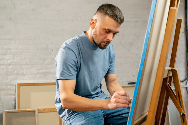 jovem artista em uma camiseta azul em um estúdio de arte trabalhando em uma pintura enquanto sentado - Foto, Imagem