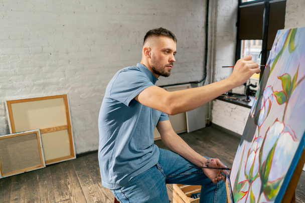 jeune artiste en t-shirt bleu dans un atelier d'art travaillant sur une peinture assis - Photo, image