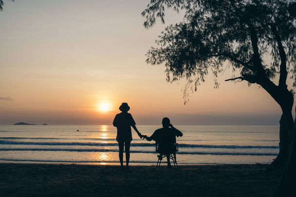Туристическая пара на пляже под деревом и наблюдать за восходом солнца, красочный золотой утренний свет, концепция наружного образа жизни. - Фото, изображение
