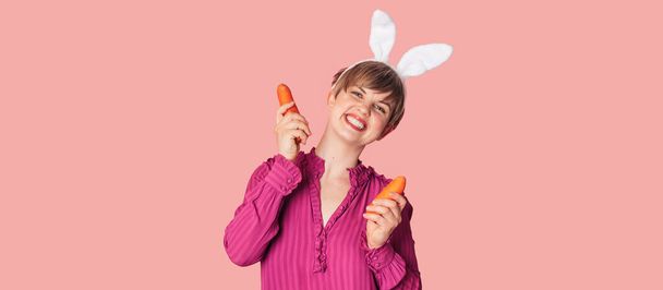 Sonriente chica caucásica delgada en orejas de conejo con zanahorias frescas en sus manos. Banner panorámico, espacio libre. Hortalizas ecológicas - Foto, imagen