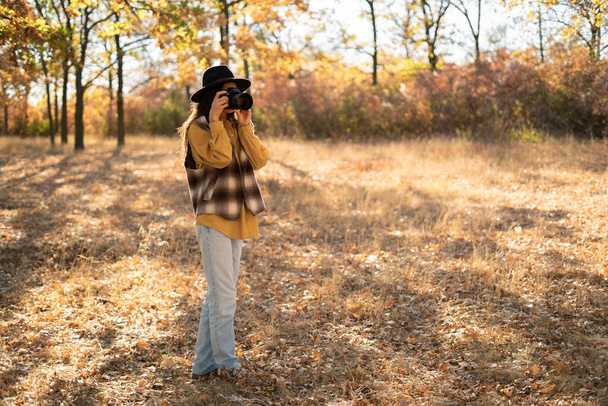 Leuke vrouw met zwarte hoed met camera geniet van zonnig weer in het najaarspark, fotograaf in het gele bos maakt foto 's. Herfst levensstijl. Zijaanzicht. Kopieerruimte - Foto, afbeelding