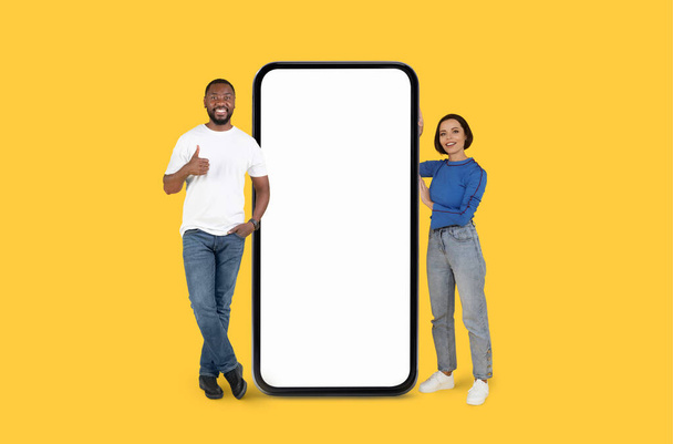 A happy black man and woman stand beside a large, blank smartphone screen on a yellow background, ideal for advertising content - Photo, Image