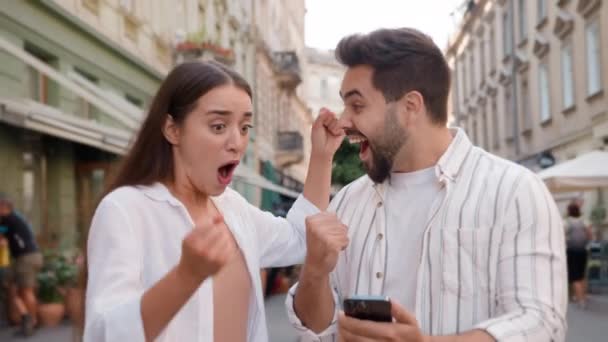 Caucasian happy couple enjoy excited man woman shocked lucky success victory mobile phone smartphone reading message good news amazed surprised open mouth city outdoors urban gen z win wonder awesome - Footage, Video