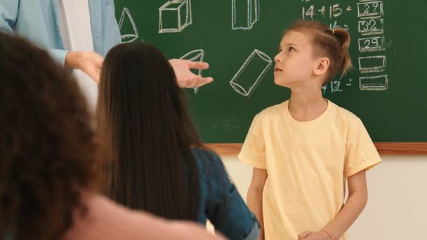 Professional teacher talking and explain idea to asian child during class. Caucasian student listening instructor while standing in front of classroom with blackboard math lesson written. Pedagogy. - Photo, Image