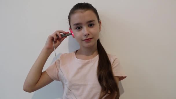 Una piccola bella bruna felice ragazza caucasica muove un bastone con una luce a infrarossi dall'orecchio sinistro a destra, trattando e riscaldandosi per un raffreddore, seduta vicino a un muro bianco in un bambino - Filmati, video