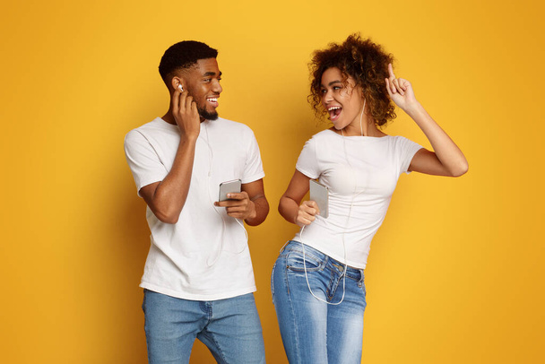 Radostné tisícileté černý muž a žena v sluchátka poslouchat hudbu na mobily a usmívá se navzájem, oranžové pozadí - Fotografie, Obrázek