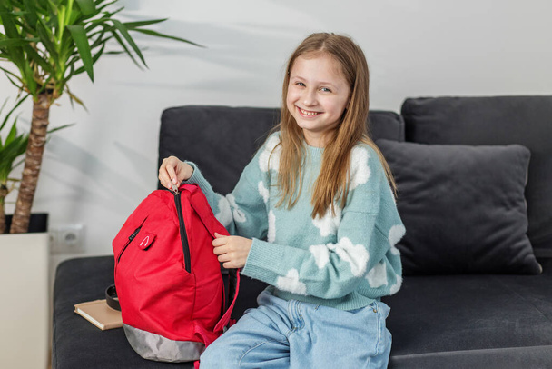 Vidám kiskorú gyermek mosolyog, miközben a csomagolás piros hátizsák, kényelmesen ül a sötét kanapén otthon. - Fotó, kép
