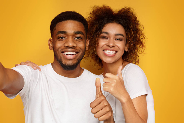 Láska je skvělá. Veselý afroamerické muž a žena dohromady takže selfie, zobrazeno thumbs up na kameru, oranžové pozadí - Fotografie, Obrázek